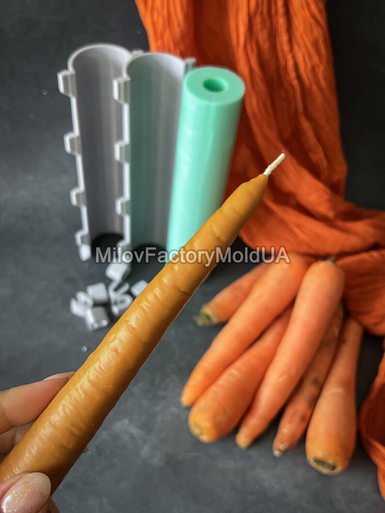 May include: A carrot-shaped candle with a white wick is held in front of a grey surface. Several orange carrots and candle moulds are also visible. The moulds are white and mint green. The background includes orange fabric.