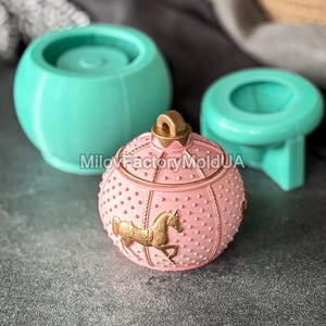 Könnte beinhalten: Ein rosa, runder Behälter mit einem goldenen Pferdemotiv und einem goldenen Griff oben. Im Hintergrund befinden sich zwei türkis farbene Formen. Der Behälter hat eine gepunktete Textur.