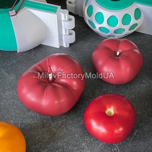 May include: Four red tomatoes and one orange tomato on a gray surface. Two white and green plastic molds are also on the surface. One mold is a sphere with holes and the other is a half sphere with a red rubber band.