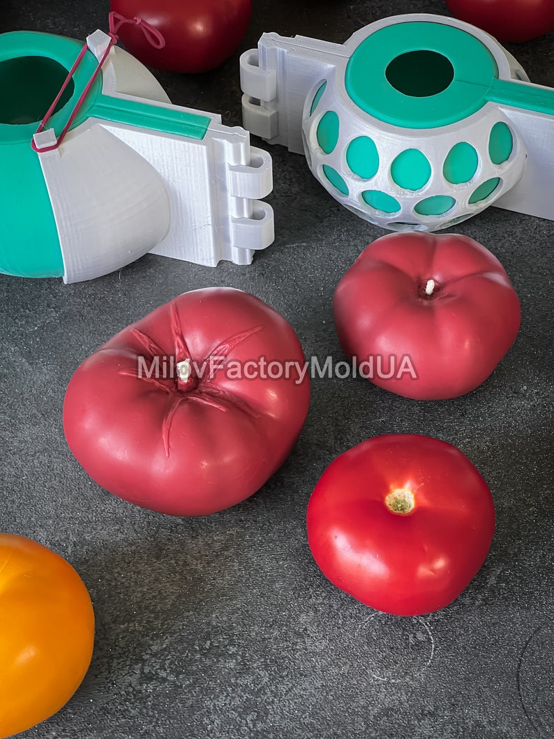 May include: Four red tomatoes and one orange tomato on a gray surface. Two white and green plastic molds are also on the surface. One mold is a sphere with holes and the other is a half sphere with a red rubber band.