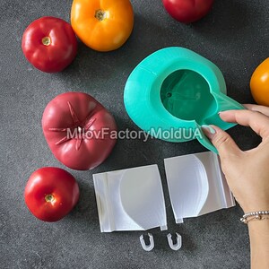 May include: A green silicone mold for making tomato-shaped objects. The mold is open and a hand is holding the top half of the mold. There are several red tomatoes and one orange tomato on a gray surface. The mold is labeled "MiloFactoryMoldUA".
