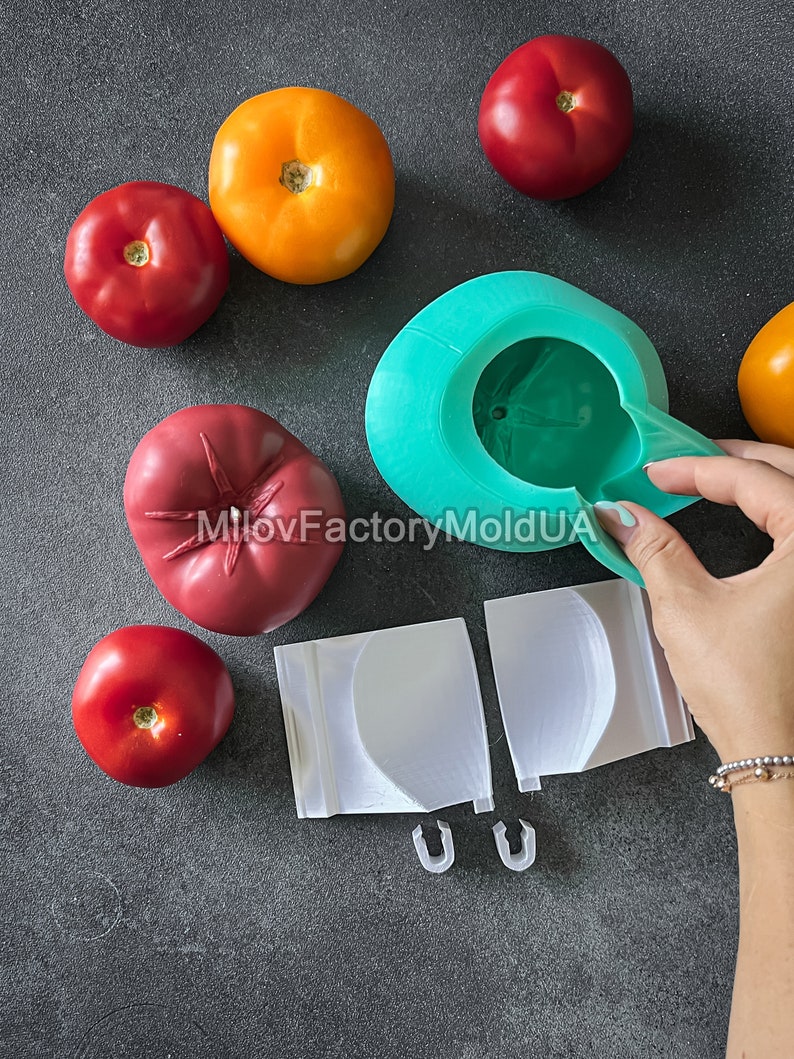 May include: A green silicone mold for making tomato-shaped objects. The mold is open and a hand is holding the top half of the mold. There are several red tomatoes and one orange tomato on a gray surface. The mold is labeled "MiloFactoryMoldUA".