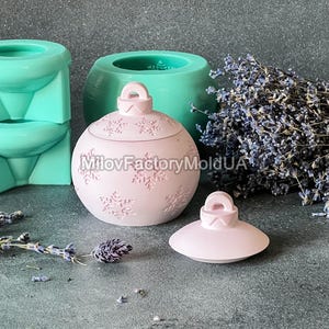 Pu&ograve; includere: Un ornamento natalizio in ceramica rosa con un motivo a fiocco di neve e un coperchio. L'ornamento &egrave; su una superficie grigia con un mazzo di fiori di lavanda sullo sfondo.