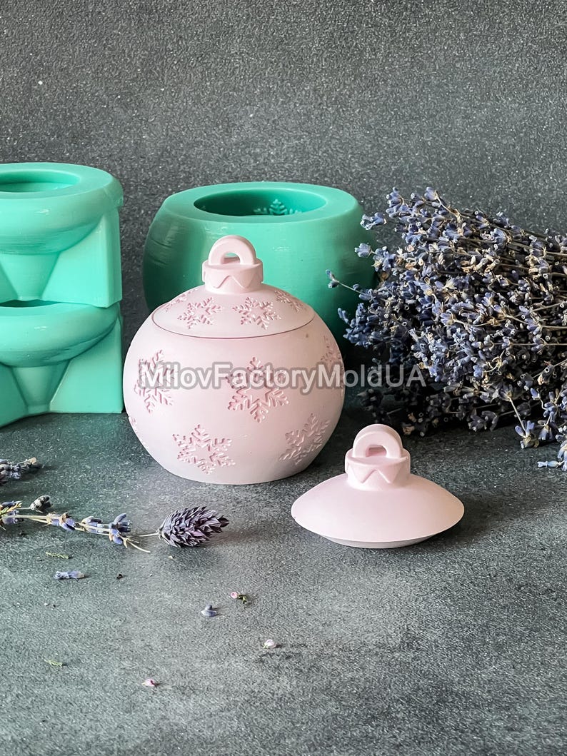 Pu&ograve; includere: Un ornamento natalizio in ceramica rosa con un motivo a fiocco di neve e un coperchio. L'ornamento &egrave; su una superficie grigia con un mazzo di fiori di lavanda sullo sfondo.
