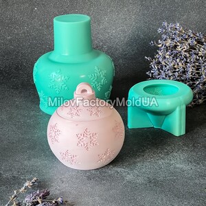 Pu&ograve; includere: Tre stampi in silicone per realizzare candele a forma di ornamento natalizio. Gli stampi sono turchese e rosa e presentano un motivo a fiocco di neve. Gli stampi sono su una superficie grigia.