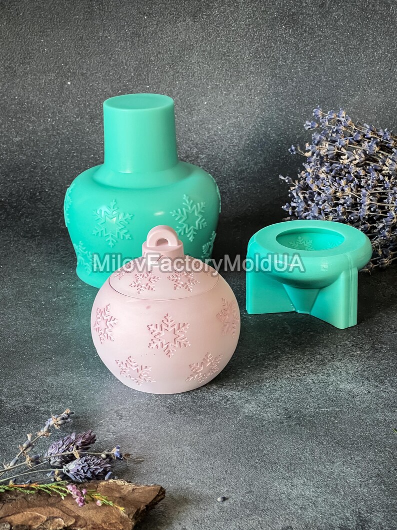 Pu&ograve; includere: Tre stampi in silicone per realizzare candele a forma di ornamento natalizio. Gli stampi sono turchese e rosa e presentano un motivo a fiocco di neve. Gli stampi sono su una superficie grigia.