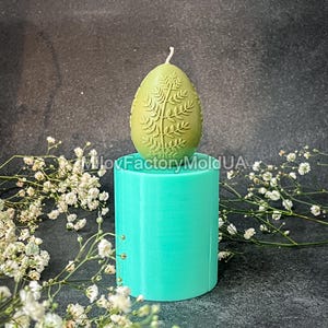 May include: A teal silicone mould for making a candle in the shape of an egg with a leafy design. The mould is sitting on a black surface with white flowers.