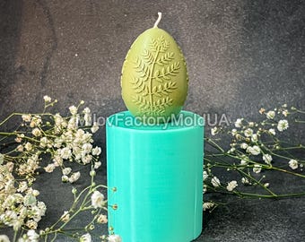 May include: A teal silicone mold for making a candle in the shape of an egg with a leafy design. The mold is sitting on a black surface with white flowers.