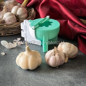 Pode incluir: Uma forma de silicone verde para fazer velas em forma de alho. A forma está aberta e mostra o interior da forma. Há três bulbos de alho inteiros e uma vela em forma de alho sobre uma superfície cinza.