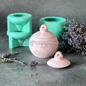 Pu&ograve; includere: Un ornamento natalizio rosa con un motivo a fiocco di neve e un coperchio. L'ornamento &egrave; in resina e si trova su una superficie grigia. L'ornamento &egrave; accanto a uno stampo in silicone verde.