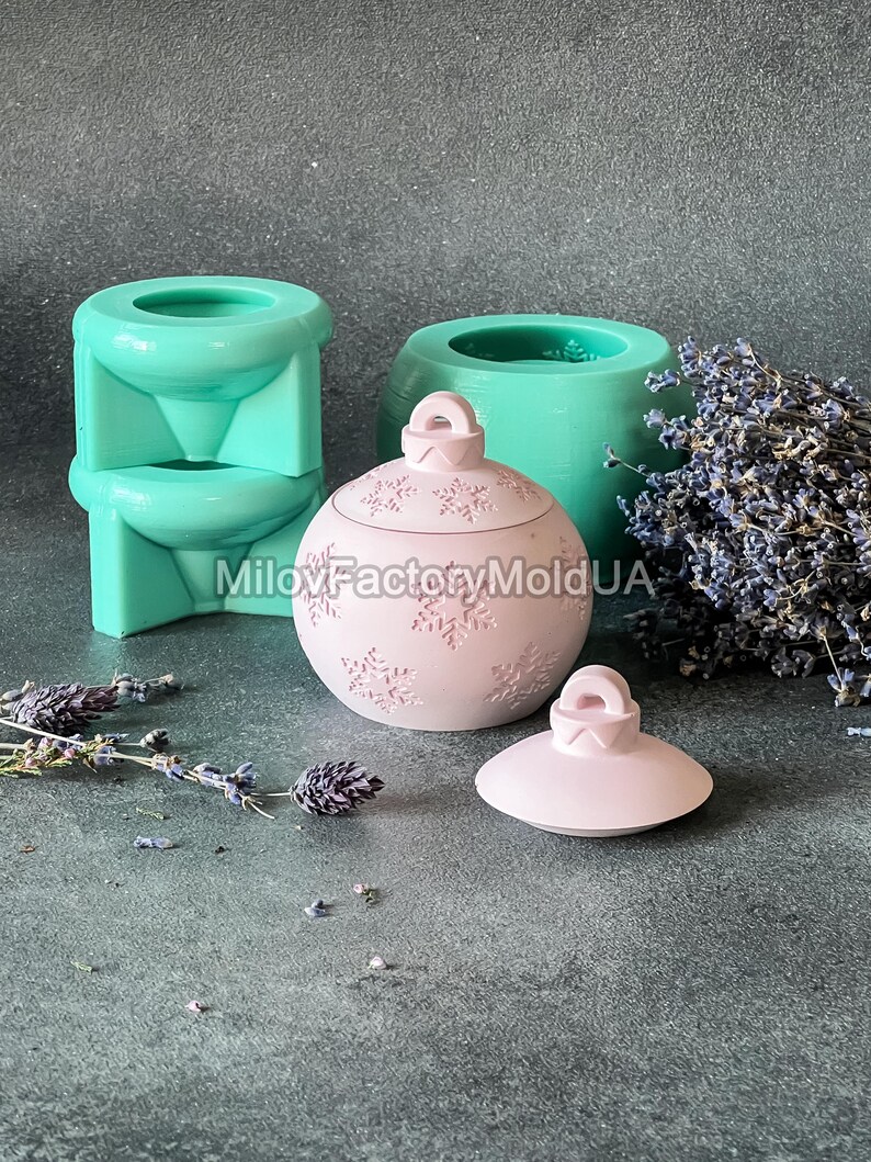 Pu&ograve; includere: Un ornamento natalizio rosa con un motivo a fiocco di neve e un coperchio. L'ornamento &egrave; in resina e si trova su una superficie grigia. L'ornamento &egrave; accanto a uno stampo in silicone verde.