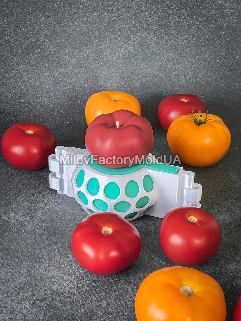 May include: A red tomato candle sits on top of a white and teal 3D printed mold with six whole tomatoes surrounding it. The tomatoes are a mix of red and orange.
