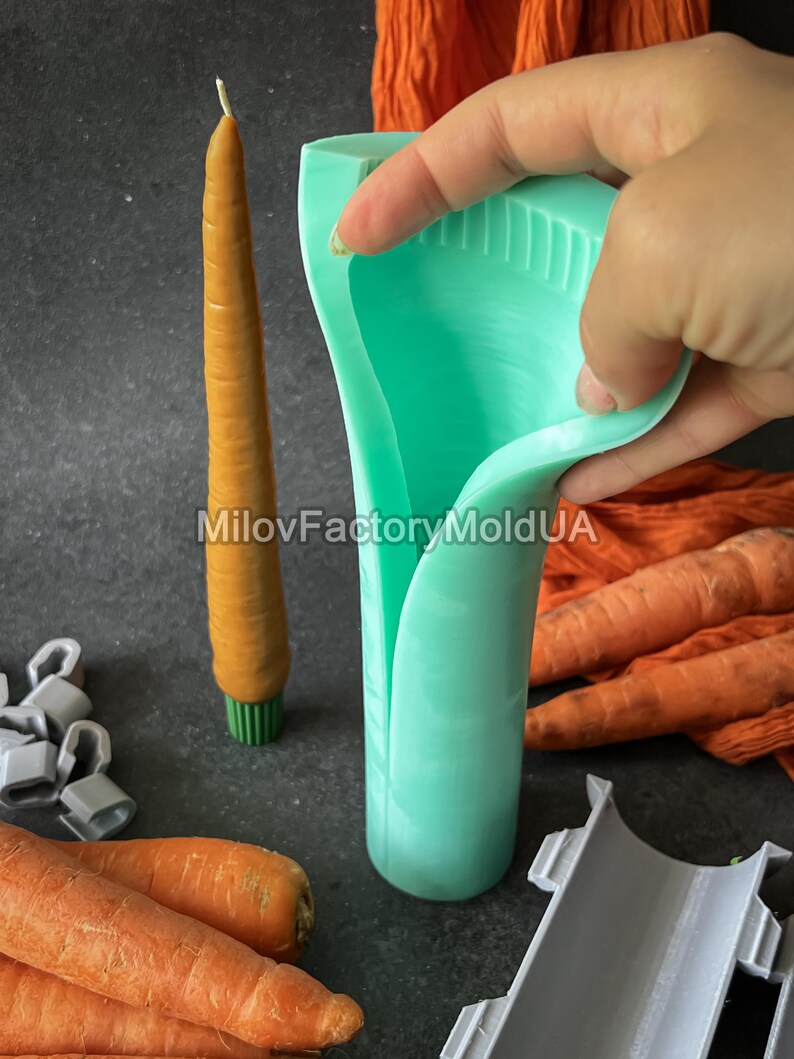 May include: A light green silicone carrot-shaped mould is held in a hand, next to a carrot-shaped candle and fresh carrots. The candle is orange with a green base. The image includes the text "MilovFactoryMoldUA".