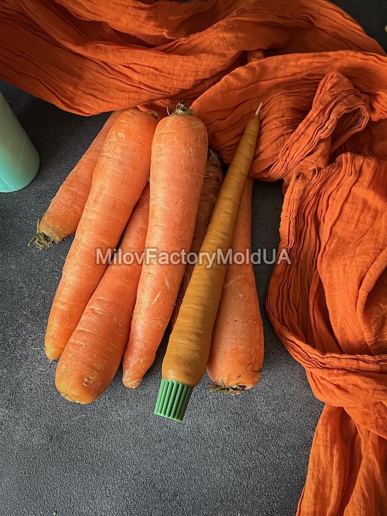 May include: Several fresh, orange carrots with green tops are arranged with a carrot-shaped candle with a green brush base. An orange fabric is draped in the background. The text "MilovFactoryMoldUA" is visible.