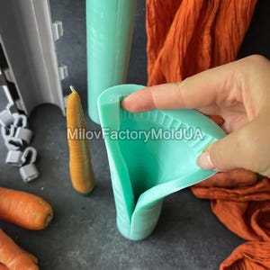 May include: A teal silicone mould for making carrot-shaped candles is held open, revealing the interior. A finished carrot candle and other moulds are visible. Orange fabric and fresh carrots are also in the image. The text "MilovFactoryMoldUA" is visible.