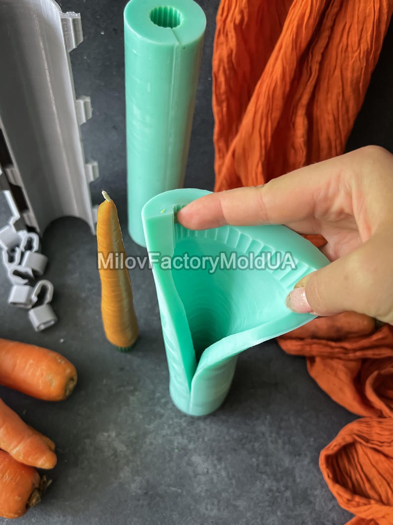 May include: A teal silicone mould for making carrot-shaped candles is held open, revealing the interior. A finished carrot candle and other moulds are visible. Orange fabric and fresh carrots are also in the image. The text "MilovFactoryMoldUA" is visible.