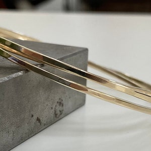 以下が含まれることがあります： 3つの金のバングルが積み重ねられています。バングルは滑らかで、わずかにハンマーで叩かれたような質感があります。