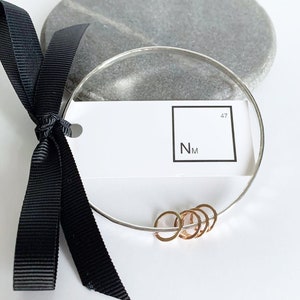 May include: A silver bangle bracelet with a hammered finish and five small gold rings attached. The bracelet is displayed on a gray stone surface with a white tag that reads "NM 47".