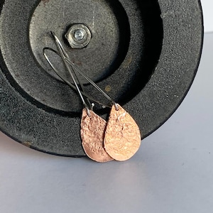 May include: A pair of copper teardrop earrings with a hammered texture. The earrings are hanging from silver hooks.