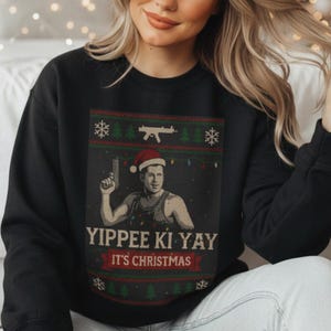 May include: Black sweatshirt with a festive design. The graphic features a man in a Santa hat holding a gun, with the text "Yippee Ki Yay It's Christmas". The design includes snowflakes, Christmas trees, and a machine gun.
