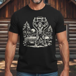 Op de afbeelding: Zwart t-shirt met een Bigfoot-afbeelding die een boombox boven zijn hoofd houdt. Het ontwerp bevat een auto, bomen en een ondergaande zon, allemaal in wit en grijs. Een leuke, originele tee.