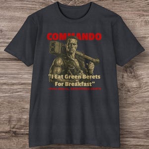 May include: Dark grey t-shirt with a graphic of a man holding a weapon, and the word "COMMANDO" in red. The quote "I Eat Green Berets For Breakfast" is below the image.