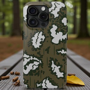 May include: A mobile phone case featuring a camouflage design in green and white. The case has a shiny finish and a raised edge around the camera lenses. The phone case is on a wooden surface.