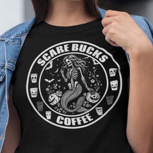 May include: A black t-shirt featuring a white circular graphic with the text SCARE BUCKS COFFEE. The design depicts a skeleton mermaid surrounded by bats, flowers, and pumpkins, with small icons of coffee cups with faces around the border.