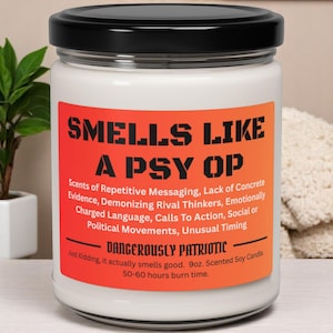 May include: A white candle in a glass jar with a black lid. The label features the text "SMELLS LIKE A PSY OP" in bold, orange letters. Additional text describes the candle's scent and burn time.