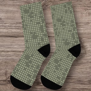 May include: A pair of olive green socks with a repeating square and dot pattern. The socks have black heels and toes. The socks are displayed on a wooden surface.