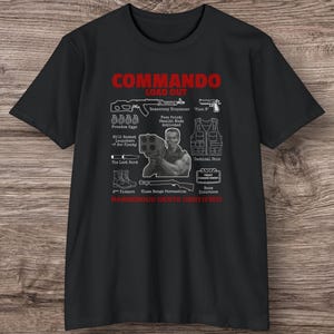 Puede incluir: Camiseta negra con un gráfico rojo que dice "COMMANDO LOAD OUT". El gráfico incluye ilustraciones de armas y el texto "DANGEROUS GENTS CERTIFIED".