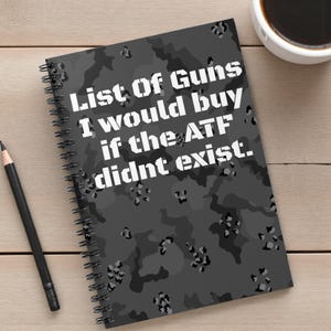 Puede incluir: Un cuaderno con espiral con un patrón de camuflaje gris y el texto "List of Guns I would buy if the ATF didnt exist." Un lápiz negro y una taza de café también son visibles.