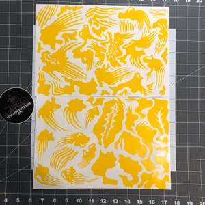 Puede incluir: Dos hojas de pegatinas de vinilo amarillas con diseños abstractos. Las pegatinas presentan varias formas, incluyendo formas de nubes y elementos lineales. Un logotipo en blanco y negro con texto es visible en la esquina inferior izquierda.