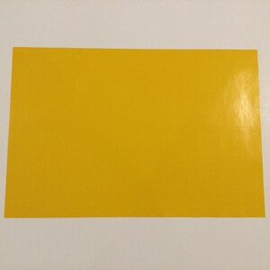 May include: A rectangular sheet of bright yellow paper, centered on a white surface. The paper has a glossy finish and appears to be a solid color with no visible patterns or text. The edges are clean and well-defined.