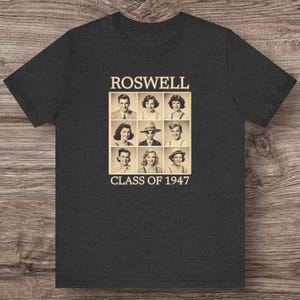 May include: Dark grey t-shirt with a vintage graphic. The graphic features the word "ROSWELL" above a grid of nine sepia-toned portraits and the text "CLASS OF 1947" below. The shirt is made of a soft material.