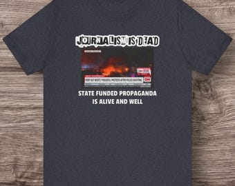 May include: Dark gray t-shirt with the text "JOURNALISM IS DEAD" above a news report image and the text "STATE FUNDED PROPAGANDA IS ALIVE AND WELL". The news report image shows a fiery scene with the CNN logo.