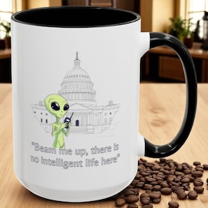 May include: White ceramic mug with a black handle and rim. The mug features a cartoon alien holding a device in front of a Capitol building illustration. Text on the mug reads: "Beam me up, there is no intelligent life here."