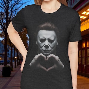 Könnte beinhalten: Dunkelgraues T-Shirt mit einem Schwarz-Weiß-Bild eines Gesichts mit weißer Maske und Händen, die eine Herzform bilden. Das T-Shirt hat einen Rundhalsausschnitt.
