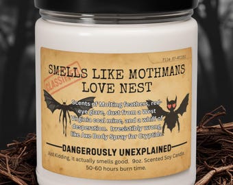 May include: A white scented soy candle in a glass jar with a black lid. The label reads "Smells Like Mothmans Love Nest" with illustrations of a mothman and bat. The candle has a 9oz volume and a 50-60 hour burn time.