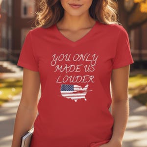 Pode incluir: Camiseta vermelha com decote em V com o texto branco "You Only Made Us Louder". O design inclui uma bandeira americana dentro do contorno dos Estados Unidos.