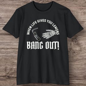 May include: Black t-shirt with white text and graphics. The text reads "WHEN LIFE GIVES YOU LEMONS" above a graphic of a hand skeleton and a small object. Below the text is the phrase "BANG OUT!"