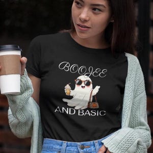 May include: A black short-sleeved t-shirt featuring a graphic of a white ghost wearing sunglasses, holding a coffee cup, and carrying a brown patterned handbag. The text BOOJEE AND BASIC is printed in white above and below the ghost illustration.