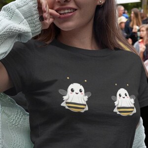 May include: A black short-sleeved t-shirt featuring two identical graphic illustrations of a small white ghost wearing a yellow and black striped bee costume. The ghosts have round black eyes, pink cheeks, thin antennae with yellow tips, and small wings.