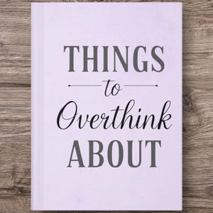 May include: A lilac-coloured journal with the words "THINGS to Overthink ABOUT" in grey and black lettering. The journal is closed and resting on a wooden surface.
