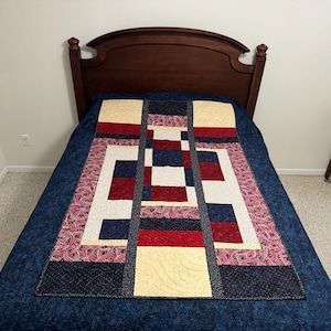 May include: A blue, red, and cream patchwork quilt with a geometric design. The quilt is on a blue bed with a wooden headboard.