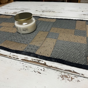 May include: A black and brown quilted table runner with a white candle in a glass jar on top. The candle jar has a gold lid and the word "Ginger" printed on the label.