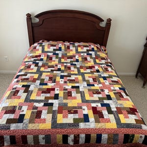 May include: A patchwork quilt with a geometric pattern in shades of red, yellow, brown, and blue. The quilt is on a bed with a wooden headboard.