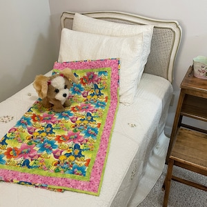May include: A white bed with a white and pink floral patterned quilt. A stuffed dog toy is lying on the quilt. A wooden nightstand with a small decorative basket is next to the bed.