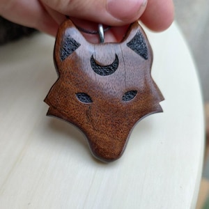 Könnte beinhalten: Eine Holz-Anhänger-Halskette mit einem geschnitzten Wolfskopf-Design. Der Anhänger hat eine satte braune Farbe mit schwarzen Akzenten in den Augen, Ohren und einem Halbmond über dem Kopf. Die Halskette ist an einer dunklen Kordel befestigt.
