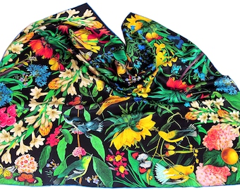 Puede incluir: Un pañuelo de seda negro con un vibrante diseño floral y de pájaros. El pañuelo presenta flores coloridas en tonos rosa, amarillo y azul, junto con follaje verde y varios pájaros y mariposas. El pañuelo tiene un borde azul.